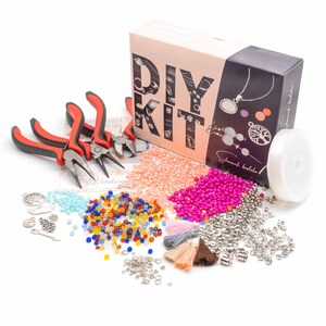 Może przedstawiać: Zestaw do samodzielnego tworzenia biżuterii DIY z różnymi koralikami, zawieszkami i narzędziami. Zestaw zawiera pudełko z napisem "DIY Kit", szpulkę nici i parę szczypiec. Korale są w różnych kolorach, w tym różowym, brzoskwiniowym, niebieskim i żółtym. Zawieszki obejmują drzewo, koło i serce. Zestaw idealnie nadaje się do tworzenia naszyjników, bransoletek i kolczyków.