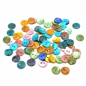 Op de afbeelding: Een verzameling ronde knopen met twee gaten in diverse iriserende kleuren, waaronder blauw, groen, geel, roze en oranje. Elke knoop heeft een glad, gepolijst oppervlak met een natuurlijk, wervelend patroon dat kenmerkend is voor schelpmateriaal.