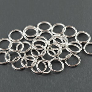 May include: A collection of small, silver-colored jump rings scattered on a dark gray surface. The rings are circular with a small opening, suggesting they are for jewelry making or crafts. The lighting highlights their metallic sheen.