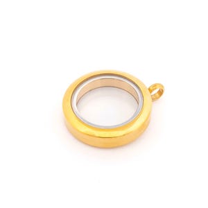 Pode incluir: Um pingente de medalhão redondo dourado com um centro de vidro transparente e um anel interno prateado. O medalhão tem uma pequena argola para prender a uma corrente. O pingente está sobre um fundo branco.
