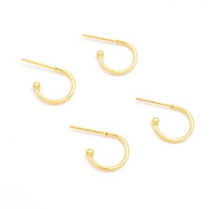 Peut inclure: Quatre boucles d'oreilles dorées en forme de cercle, avec une petite boule à l'extrémité. Les boucles d'oreilles sont sur fond blanc. Elles sont petites et délicates.
