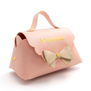 Peut inclure: Un sac à main rose avec une poignée et un nœud. Le sac a un dessus festonné et les mots "SWEET DAY" en lettres dorées. Un bouton doré est au centre du nœud.