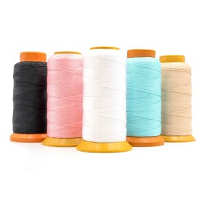 Könnte beinhalten: Fünf Garnrollen in verschiedenen Farben: Schwarz, Rosa, Weiß, Hellblau und Beige. Jede Rolle hat eine gelbe Basis und einen orangefarbenen Deckel. Das Garn ist eng gewickelt und erzeugt ein strukturiertes Aussehen.
