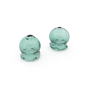 May include: Two teal glass decorative objects. Each has a rounded top with a small opening and a base with a unique, curved design. The glass is slightly transparent. The objects are set against a white background. The objects are likely for home decor.