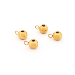 May include: Four small, gold-coloured metal beads, each with a loop. The beads are spherical with a small hole at the top. The beads are on a white background.