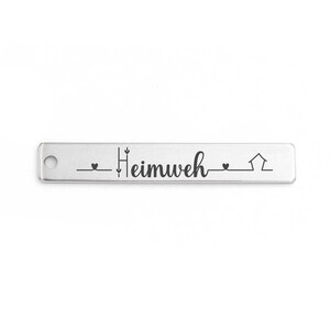 Könnte beinhalten: Ein rechteckiger Anhänger aus silberfarbenem Metall mit einem kleinen runden Loch auf der linken Seite. Die Oberfläche ist mit dem Wort "Heimweh" in Schreibschrift graviert, flankiert von zwei kleinen Herzsymbolen und einer Strichzeichnung eines Hauses.