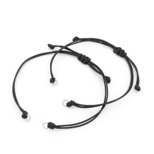 Pode incluir: Duas pulseiras de cordão preto com nós deslizantes ajustáveis e pequenos anéis de metal prateado nas extremidades dos cordões. As pulseiras são feitas de um fio preto fino e liso, concebidas com uma estrutura circular simples.