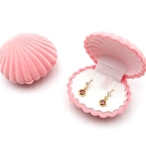 Könnte beinhalten: Eine rosa muschelförmige Schmuckschatulle, geöffnet, um ein Paar goldene Ohrringe mit rosa Edelsteinen zu zeigen. Die Schatulle besteht aus einem weichen, samtigen Material. Die Ohrringe werden auf weißem Hintergrund präsentiert.