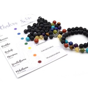 Pode incluir: Um bracelete de pedra de lava preta com sete pedras de chakra. As pedras são jaspe vermelho, olho de tigre, citrino, aventurina verde, lapis-lazúli, ametista e quartzo claro. O bracelete está sobre um fundo branco com um cartão que lista os sete chakras e suas pedras correspondentes.