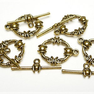 Könnte beinhalten: Goldfarbene Metall-Schiebeverschlüsse mit verzierten Designs und runden Akzenten. Die Verschlüsse haben eine Stange, die durch eine Schlaufe gleitet, um zu sichern.