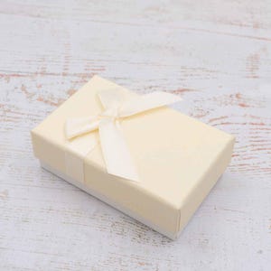 Op de afbeelding: Een kleine, rechthoekige geschenkdoos in een lichtgele kleur, vastgebonden met een bijpassend satijnen lint en strik. De doos heeft een getextureerd oppervlak en een witte basis, geschikt voor sieraden of kleine cadeaus.