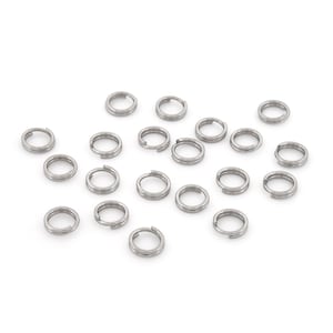 Puede incluir: Una colección de pequeños anillos partidos de color plateado, dispersos sobre una superficie blanca. Estos anillos de doble bucle son de metal y se utilizan habitualmente en la fabricación de joyas, llaveros y otros proyectos de artesanía. Vista superior.