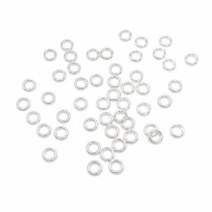May include: A large quantity of silver jump rings, scattered on a white background. The rings are small and circular, with a smooth, polished finish.