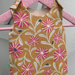 Peut inclure: Un bavoir pour bébé jaune moutarde avec un motif floral de fleurs roses et de feuilles blanches. Le bavoir a une doublure crème et est orné de petites coccinelles bleues. Il est suspendu à un cintre en bois rose.