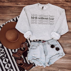 May include: A pale grey sweatshirt featuring the words "birth without fear" stacked. It's styled with light blue denim shorts, brown heeled sandals, a wide-brimmed brown hat, and sunglasses. The items are arranged on a wooden surface.