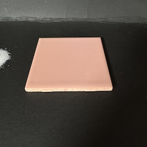 May include: A square, pale-pink ceramic tile with a smooth, glossy surface. The tile has a light-brown, textured edge. The tile is displayed on a black surface.
