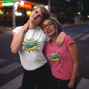 Pode incluir: Duas pessoas vestindo camisetas com um gr&aacute;fico de "OMG!" estilo quadrinhos. A pessoa &agrave; esquerda est&aacute; vestindo uma camiseta branca e a pessoa &agrave; direita est&aacute; vestindo uma camiseta rosa.