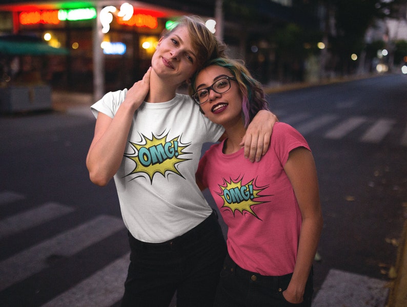 Pode incluir: Duas pessoas vestindo camisetas com um gr&aacute;fico de "OMG!" estilo quadrinhos. A pessoa &agrave; esquerda est&aacute; vestindo uma camiseta branca e a pessoa &agrave; direita est&aacute; vestindo uma camiseta rosa.