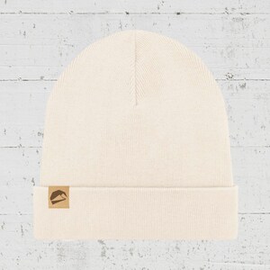 Könnte beinhalten: Eine cremefarbene Strickmütze mit einem braunen Lederpatch mit einem Berglogo.
