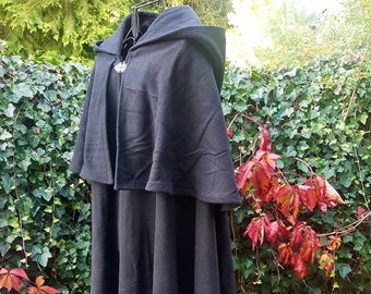 Puede incluir: Una capa larga con capucha negra con una capelina y un broche decorativo. La capa está hecha de una tela oscura y llega hasta el suelo. El fondo presenta follaje verde y hojas rojas.