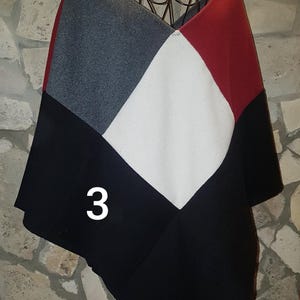 Könnte beinhalten: Ein farbblockierter Poncho in einem geometrischen Design. Der Poncho hat eine Kombination aus grauen, weißen, roten und schwarzen Paneelen. Die Zahl "3" wird in Weiß auf dem schwarzen Panel angezeigt. Das Kleidungsstück wird auf einem dekorativen Metallständer präsentiert.