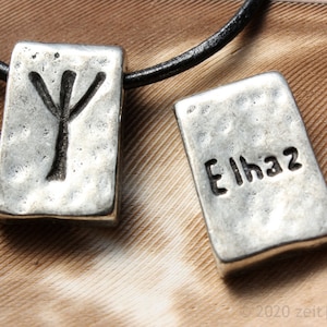 Könnte beinhalten: Zwei silberne Anhänger an einem schwarzen Lederband. Der erste Anhänger hat ein Runenzeichen, der zweite Anhänger hat das Wort "Eihwaz" eingraviert.