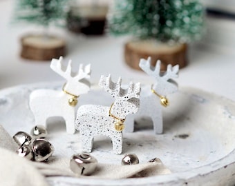 Könnte beinhalten: Drei weiße Keramik-Rentierfiguren mit goldenen Glöckchen um den Hals, die auf einem weißen Keramiktablett mit weißem Hintergrund stehen.