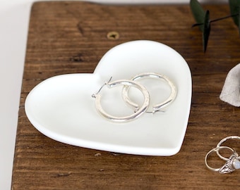 May include: A white ceramic heart-shaped dish with two silver hoop earrings inside. The dish is sitting on a wooden surface.