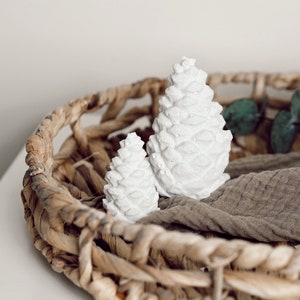 Puede incluir: Dos adornos de piña de pino de cerámica blanca en una cesta tejida con una tela marrón. Las piñas son de un diseño simple y moderno.
