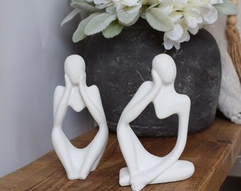 May include: Two white ceramic sculptures of abstract figures in a seated pose. The figures are stylized and have a minimalist design.