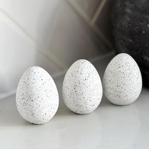 May include: Three speckled white decorative eggs. Each egg has a smooth, rounded shape and is covered in small black speckles. The eggs are arranged in a row, suggesting a minimalist or natural aesthetic.
