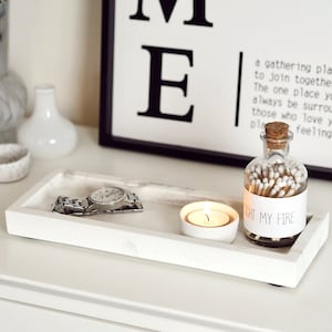 Op de afbeelding: Een wit rechthoekig dienblad met een marmerpatroon, met een zilveren horloge, een witte kaarsenhouder met een brandende kaars en een glazen fles gevuld met lucifers. De fles heeft een etiket met de tekst "Light My Fire".