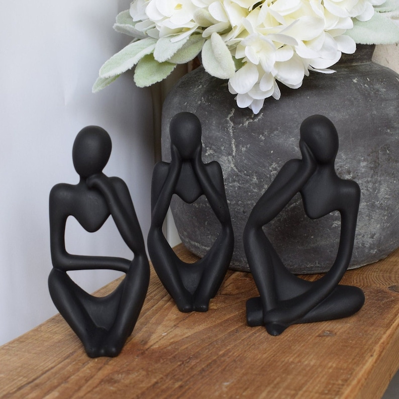 May include: Three black abstract sculptures of people sitting with their chins on their hands. The sculptures are made of a smooth, matt material and have a modern, minimalist design.