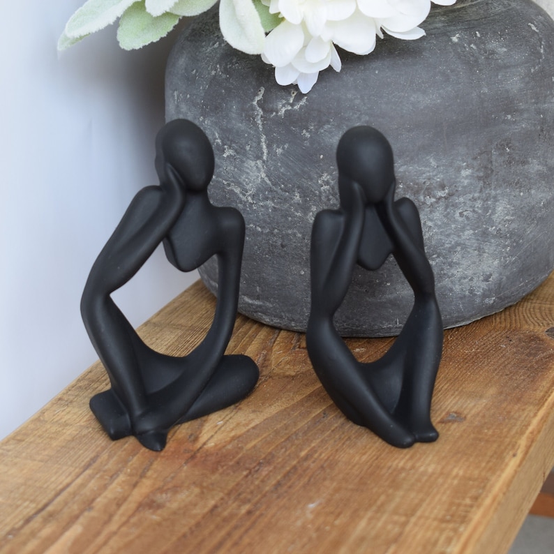 May include: Two black abstract sculptures of people sitting with their hands on their faces. The sculptures are made of a smooth, matt material and have a modern, minimalist design.