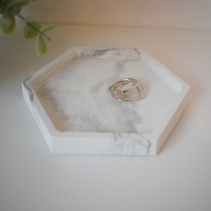 Könnte beinhalten: Ein weißes sechseckiges Tablett mit einem grauen Marmormuster, auf dem zwei silberne Ringe mit einem rosafarbenen Stein liegen. Das Tablett ist perfekt zum Aufbewahren von Schmuck oder anderen kleinen Gegenständen.