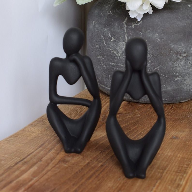 May include: Two black abstract sculptures of people sitting with their heads in their hands. The sculptures are made of a smooth, matt material and have a simple, modern design.