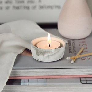 May include: A white concrete candle holder with a small, round, white candle burning inside. The candle holder is sitting on a stack of books with a book about crystals on top.