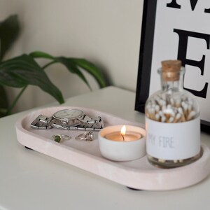 Puede incluir: Una bandeja ovalada rosa con un reloj, anillos, una peque&ntilde;a vela blanca y un frasco de vidrio lleno de cerillas. El frasco tiene una etiqueta que dice "My Fire".