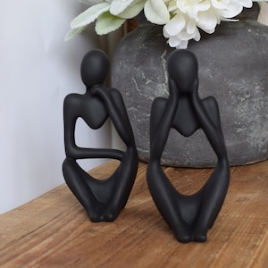 May include: Two black abstract ceramic sculptures of people sitting with their hands on their faces. The sculptures are stylised and have a minimalist design.