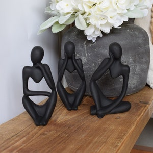 May include: Three black abstract sculptures of people sitting with their chins on their hands. The sculptures are made of a smooth, matt material and have a minimalist design.