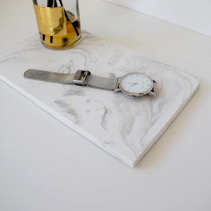Puede incluir: Una bandeja de mármol blanco con un reloj de malla plateada en la parte superior. El reloj tiene una esfera blanca y manecillas plateadas.
