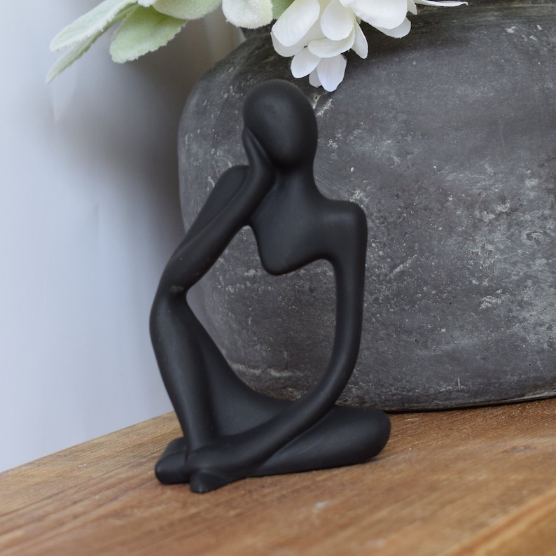 May include: Black abstract sculpture of a person sitting with their chin resting on their hand. The sculpture is made of a smooth, matt material.