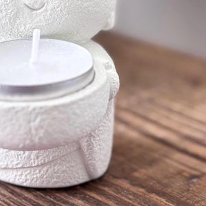 May include: A white, textured ceramic candle holder in the shape of a figure, holding a lit tea light candle. The candle holder has a smiling face and is set against a wooden surface.