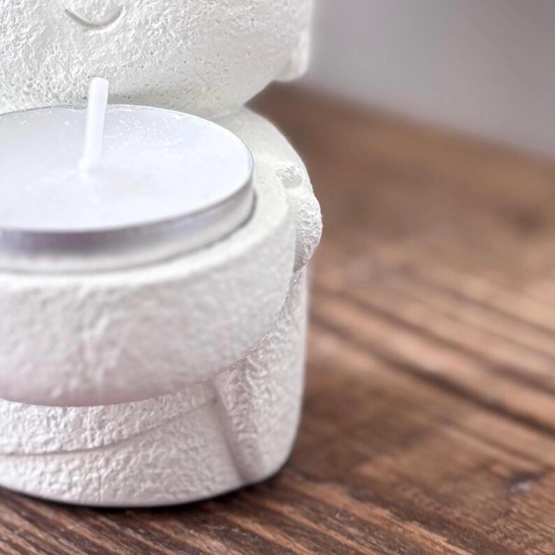 May include: A white, textured ceramic candle holder in the shape of a figure, holding a lit tea light candle. The candle holder has a smiling face and is set against a wooden surface.