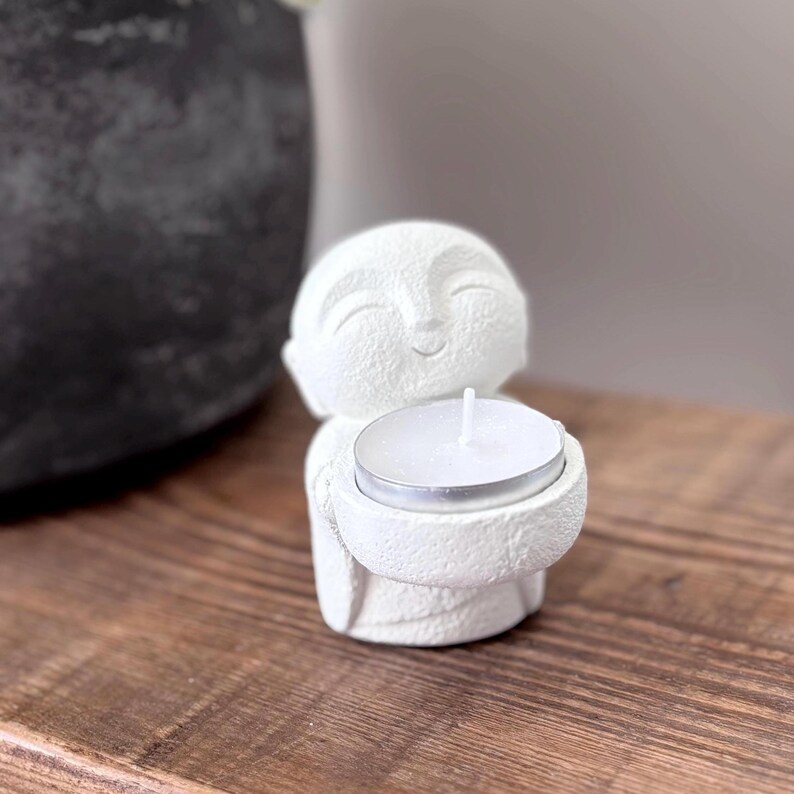 May include: A white Buddha figurine holding a small, round candle. The figurine has a smiling face and is made of a textured material. The candle is in a silver metal holder. The figurine is on a wooden surface.