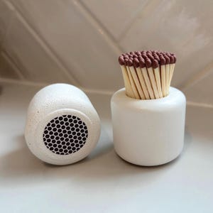 May include: A white ceramic match holder with a cylindrical shape, holding a bundle of matches with brown tips. Beside it is a white, egg-shaped object with a black honeycomb pattern.