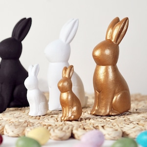 May include: Four ceramic bunny figurines in different sizes and colours. A black bunny, a white bunny, a gold bunny, and a smaller gold bunny.