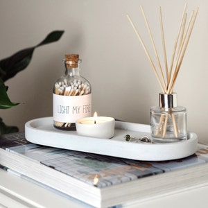Puede incluir: Una bandeja ovalada blanca con un frasco de vidrio lleno de cerillas, una peque&ntilde;a vela blanca y un difusor de vidrio con palitos de madera. El frasco de cerillas tiene una etiqueta que dice "Light My Fire".