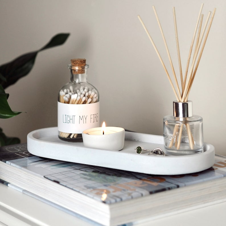 Puede incluir: Una bandeja ovalada blanca con un frasco de vidrio lleno de cerillas, una peque&ntilde;a vela blanca y un difusor de vidrio con palitos de madera. El frasco de cerillas tiene una etiqueta que dice "Light My Fire".