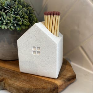 May include: A white, house-shaped match holder with a textured surface and a small square window. The holder contains several wooden matches with brown tips. The holder sits on a wooden cutting board, with a potted plant in the background.